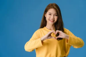an asian woman with her hands forming a heart on her chest while smiling at the camera