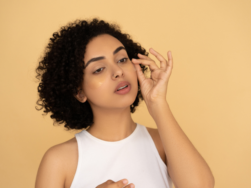 Curly-haired woman adjusting an under-eye mask against a warm background