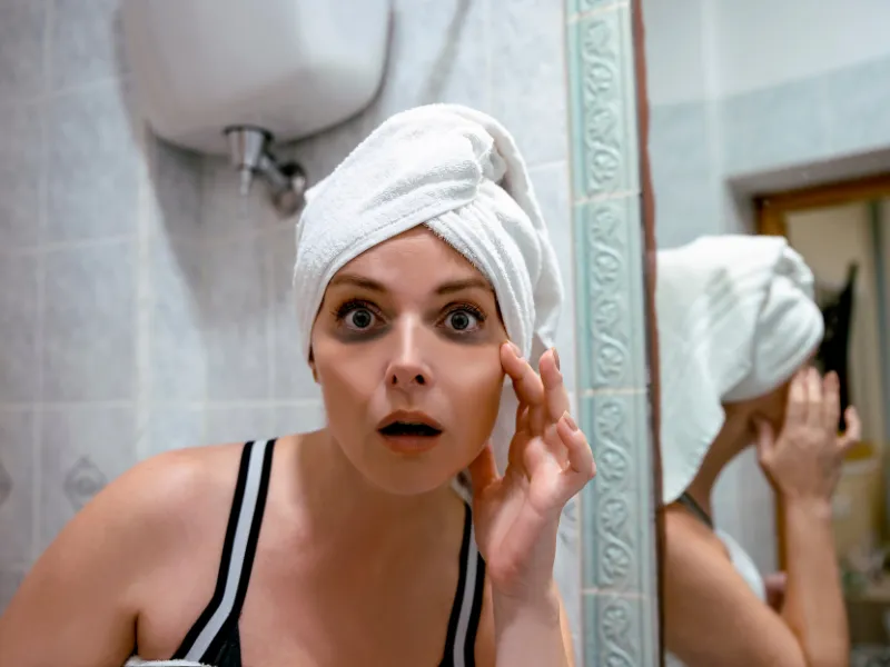 Woman with towel checking skin in bathroom mirror, looking surprised