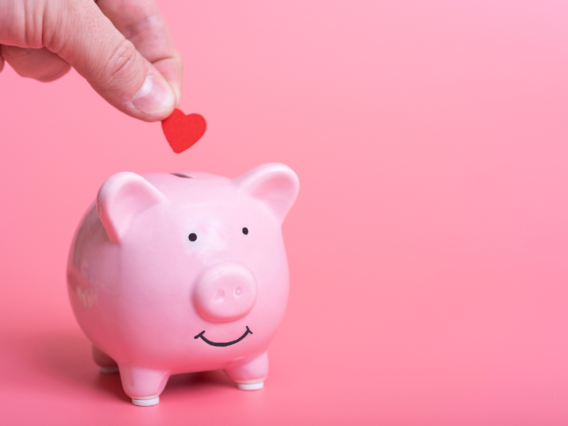 A hand putting a red heart into a pink piggy bank