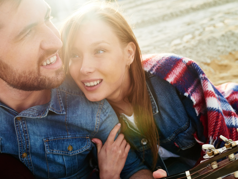 Couple cuddles on the beach wrapped in a plaid blanket