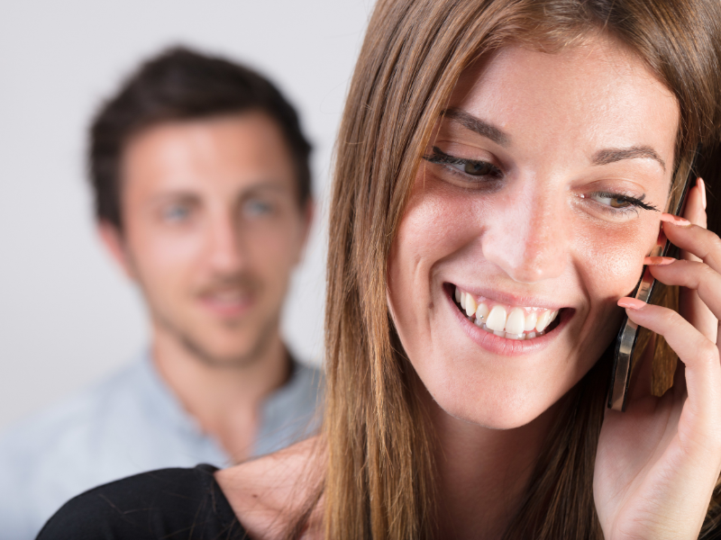 A smiling woman on phone while a man watches behind