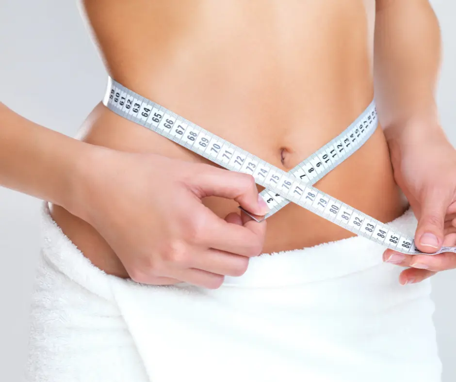 A woman measuring her waist with a tape measure while wearing a white towel