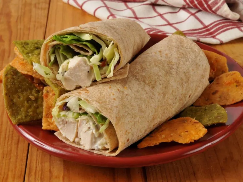 A close-up of a healthy chicken salad wrap served on a red plate with colorful tortilla chips on the side, placed on a wooden table.