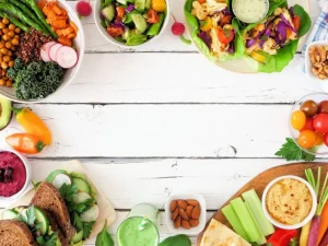 A variety of vibrant salad bowls, fresh vegetables, and healthy snacks displayed on a white wooden table, emphasizing nutritious meal options.
