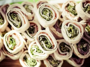 A stack of fresh vegetable wraps rolled tightly, showcasing layers of leafy greens, thinly sliced meat, and tortilla wraps