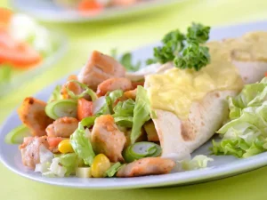A plate featuring a chicken salad wrap topped with melted cheese, accompanied by fresh lettuce and diced vegetables on a green table