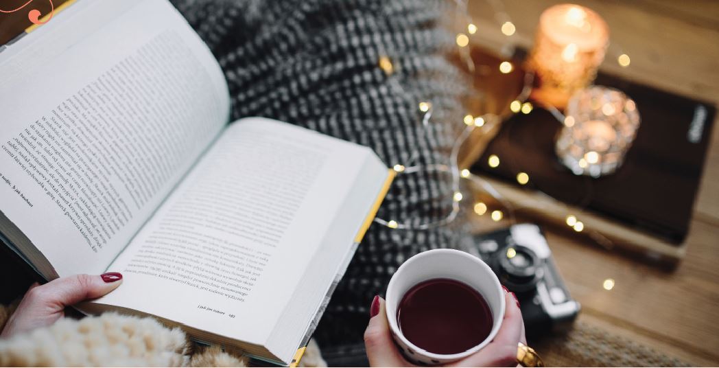 a warm and cozty picture with a warm cup of tea and candle why someone is under a blanket and reading a book