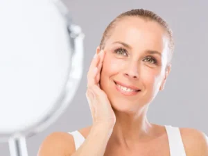 Mature woman smiling and touching her face while inspecting skin condition in a mirror